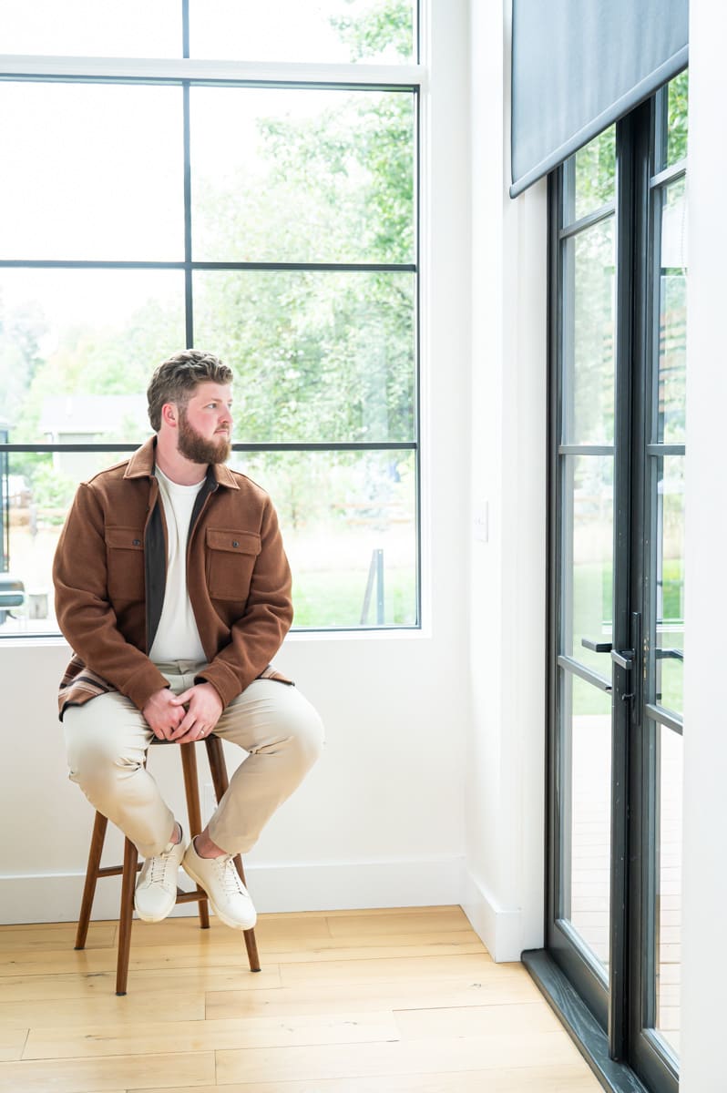 Connor Slump seated on a wooden stool beside a large black-framed window